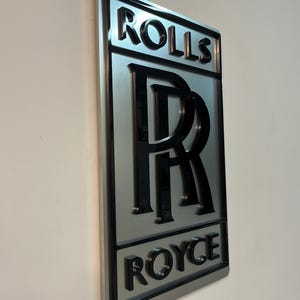 May include: A silver metal sign with a black 3D Rolls Royce logo. The logo features the iconic double R emblem and the words "Rolls" and "Royce" in a bold, black font.