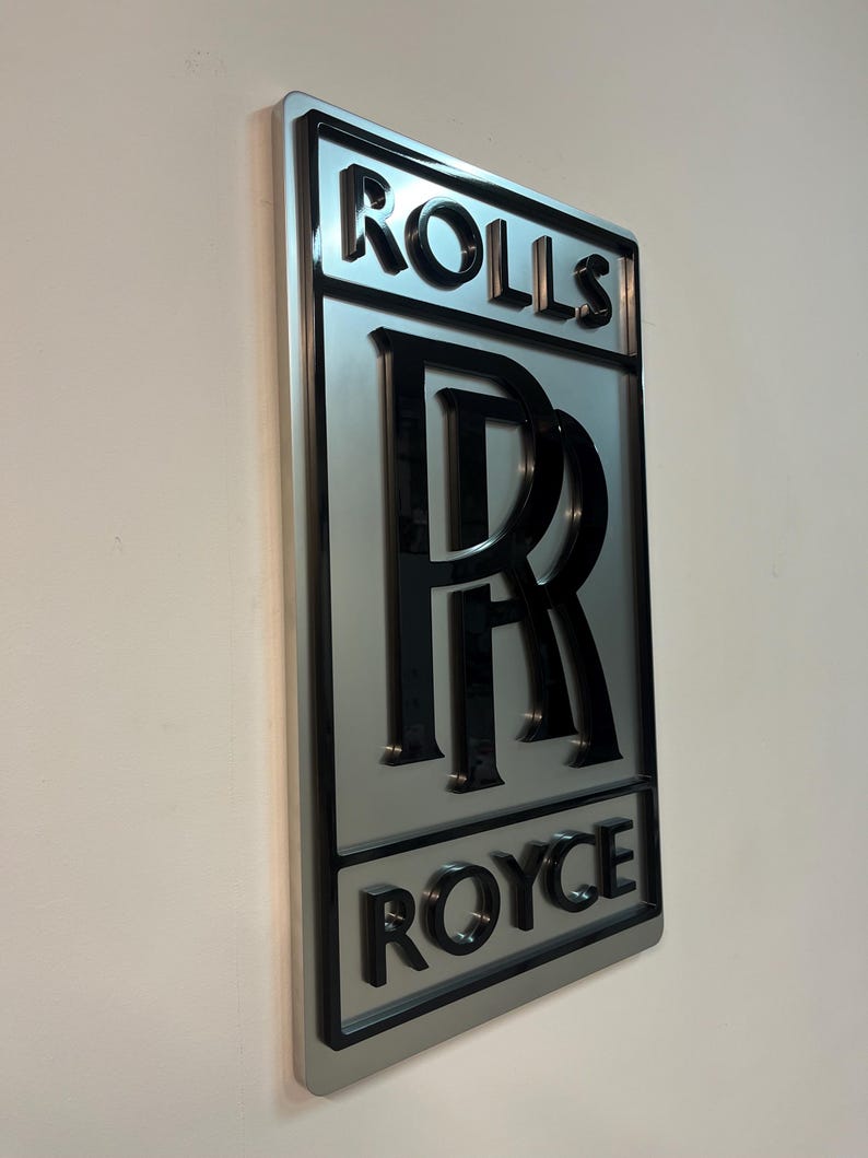 May include: A silver metal sign with a black 3D Rolls Royce logo. The logo features the iconic double R emblem and the words "Rolls" and "Royce" in a bold, black font.