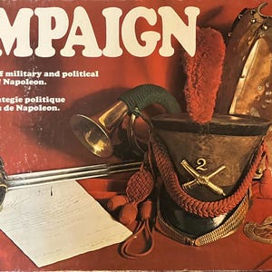 May include: The board game "Campaign" features a red background with the title in large white letters. The game includes a military helmet, sword, and armor, with text that reads "A compelling game of military and political strategy in the age of Napoleon."