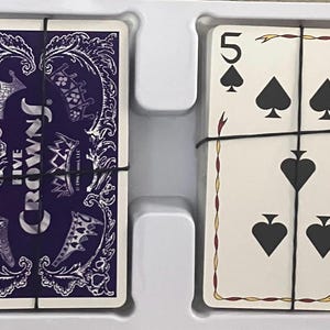Puede incluir: Un mazo de cartas Five Crowns, con un dorso morado con coronas y dise&ntilde;os ornamentados. Las cartas est&aacute;n aseguradas con una banda negra. La cara de las cartas muestra el n&uacute;mero 5 y s&iacute;mbolos de picas.