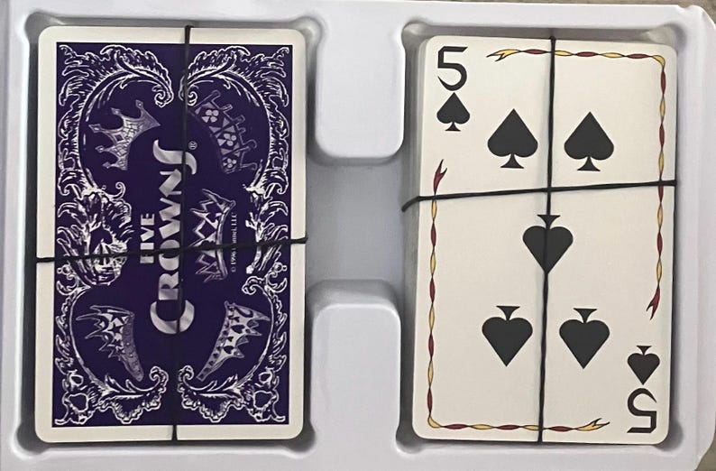 Puede incluir: Un mazo de cartas Five Crowns, con un dorso morado con coronas y dise&ntilde;os ornamentados. Las cartas est&aacute;n aseguradas con una banda negra. La cara de las cartas muestra el n&uacute;mero 5 y s&iacute;mbolos de picas.