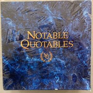 May include: A blue book with the title "Notable Quotables" in gold lettering. The book has a gold logo with the letters "NQ" inside a wreath.
