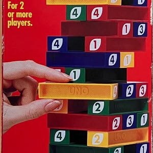 May include: A red cardboard box for the UNO STACKO game featuring a stack of colorful plastic blocks in red, yellow, green, and blue, each marked with numbers. The box displays the game title, a white UNO cube, and text including For ages 8 and older.