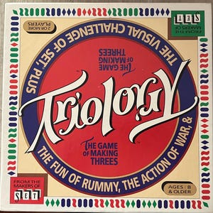 May include: A red, white, and blue game box for the game "Trilogy" by the makers of "Set". The game is described as "The Fun of Rummy, The Action of War, & The Visual Challenge of Set, Plus The Game of Making Threes".