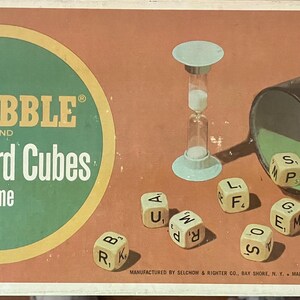 May include: A Scrabble Brand Crossword Cubes game box featuring a tan background with a green circle containing the text Scrabble Brand Crossword Cubes. The box shows a black shaker cup, a light blue hourglass, and several lettered dice cubes.