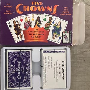 Puede incluir: Una caja del juego de cartas Five Crowns con un fondo morado y el t&iacute;tulo del juego en naranja. La caja muestra naipes con reyes y reinas. El juego es para 2-7 jugadores, a partir de 8 a&ntilde;os. Las instrucciones del juego tambi&eacute;n son visibles.