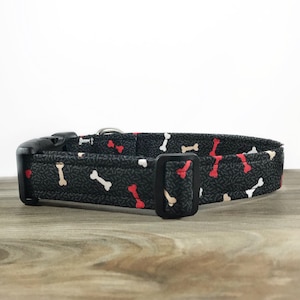 May include: Black dog collar with a bone pattern in red, white, and tan. The collar has a black buckle and a silver D-ring.
