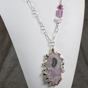 May include: A silver chain necklace with a large, irregular-shaped amethyst pendant. The pendant is set in silver and has a gold-toned edge. The necklace also features a cluster of pink and purple gemstone beads.