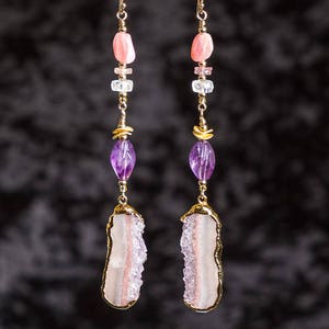 May include: A pair of long, gold earrings with a pink, white, and purple gemstone design. The earrings feature a large, white, and pink agate stone at the bottom.