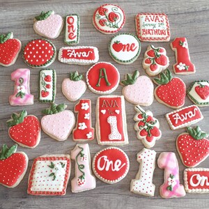 May include: A collection of decorated sugar cookies featuring strawberries, the number one, and the name Ava. The cookies are perfect for a first birthday party.