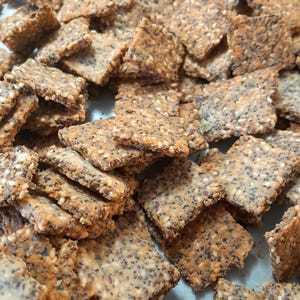 May include: A close-up of a pile of square crackers with a light brown color and a grainy texture. The crackers are covered in sesame seeds and poppy seeds.