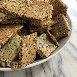 May include: A white bowl filled with a large pile of homemade crackers. The crackers are made with sesame seeds and poppy seeds.