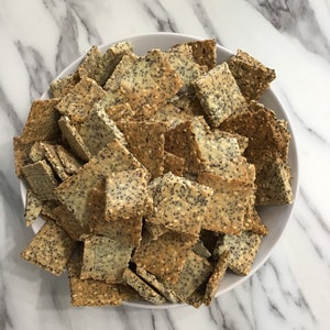 May include: A white bowl filled with a large quantity of square-shaped crackers. The crackers are covered in sesame seeds and poppy seeds.