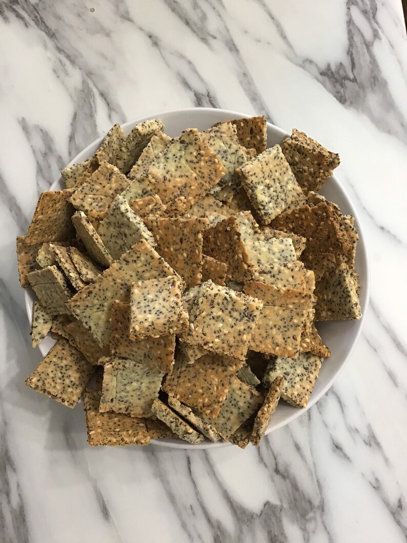 May include: A white bowl filled with a large quantity of square-shaped crackers. The crackers are covered in sesame seeds and poppy seeds.