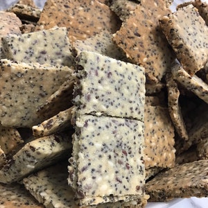May include: A close-up of a white bowl filled with a variety of crackers. The crackers are made with seeds, including sesame seeds and poppy seeds. The crackers are a light brown color and have a slightly rough texture.