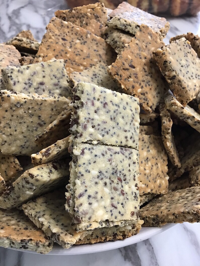 May include: A close-up of a white bowl filled with a variety of crackers. The crackers are made with seeds, including sesame seeds and poppy seeds. The crackers are a light brown color and have a slightly rough texture.