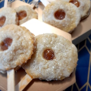 May include: Close-up of homemade mini pies with a sweet filling, topped with sugar crystals and served on wooden sticks. The pies are arranged on a brown surface with a blue and gold patterned background.