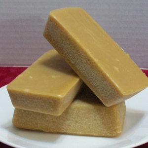 Puede incluir: Tres bloques rectangulares de fudge apilados en un plato blanco. El fudge es de color marr&oacute;n claro y tiene una superficie lisa y brillante.