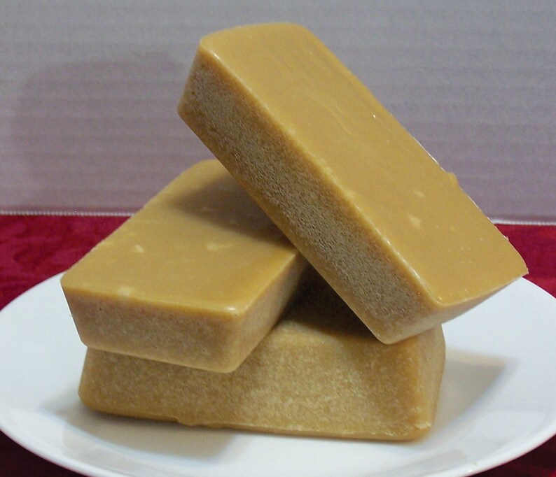 Puede incluir: Tres bloques rectangulares de fudge apilados en un plato blanco. El fudge es de color marr&oacute;n claro y tiene una superficie lisa y brillante.