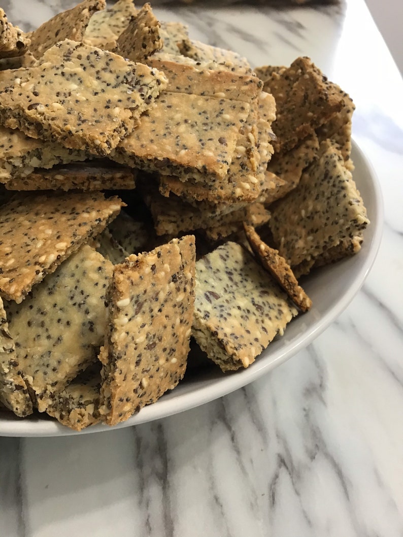 A white bowl filled with a large pile of light brown and beige crackers. The keto crackers are square and have a grainy texture. keto seed crackers by 23sweets.com