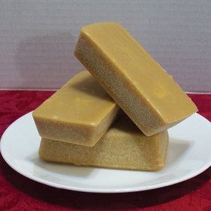 Puede incluir: Tres barras rectangulares de caramelo fudge apiladas en un plato blanco. El fudge es de color marr&oacute;n claro y tiene una superficie lisa y brillante.