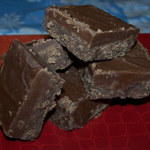 Puede incluir: Un mont&oacute;n de cuatro cuadrados de fudge de chocolate sobre una superficie roja. El fudge es marr&oacute;n oscuro y tiene una textura ligeramente granulada.
