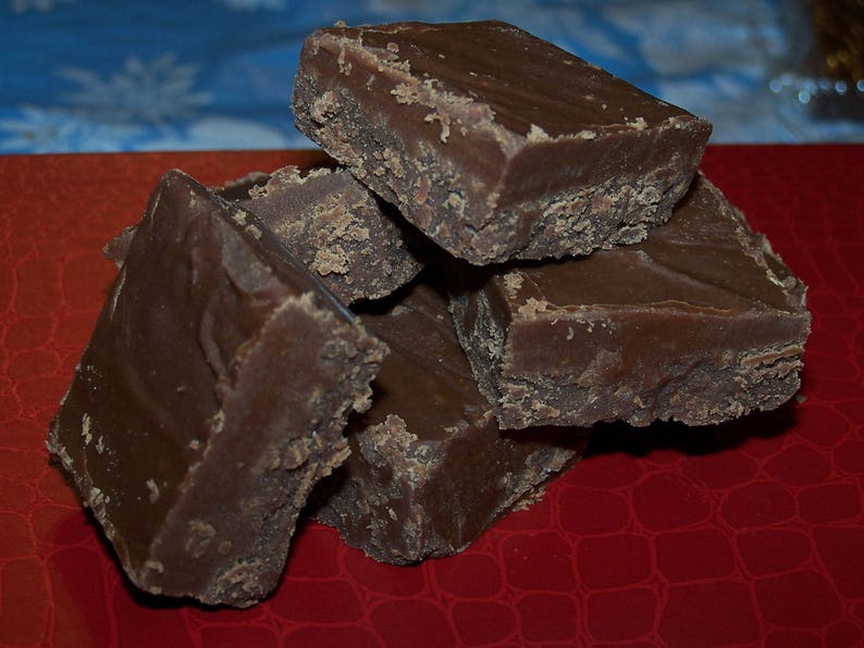 Puede incluir: Un mont&oacute;n de cuatro cuadrados de fudge de chocolate sobre una superficie roja. El fudge es marr&oacute;n oscuro y tiene una textura ligeramente granulada.