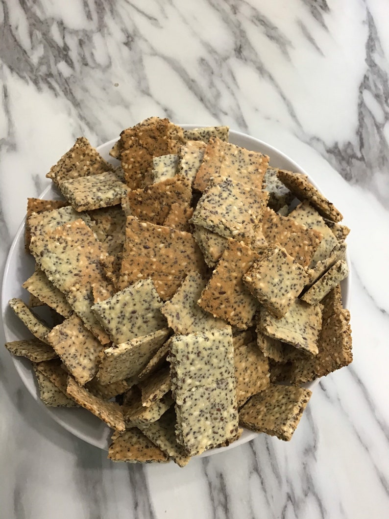 A white bowl filled with a large pile of light brown and beige crackers. The keto crackers are square and have a grainy texture. keto seed crackers by 23sweets.com