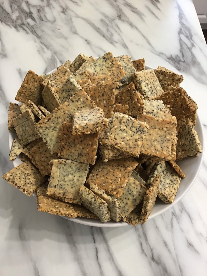 A white bowl filled with a large pile of light brown and beige crackers. The keto crackers are square and have a grainy texture. keto seed crackers by 23sweets.com