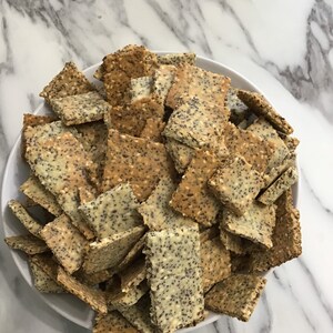 May include: A white bowl filled with a variety of crackers. The crackers are rectangular and have a light brown color with a sprinkle of poppy seeds on top.