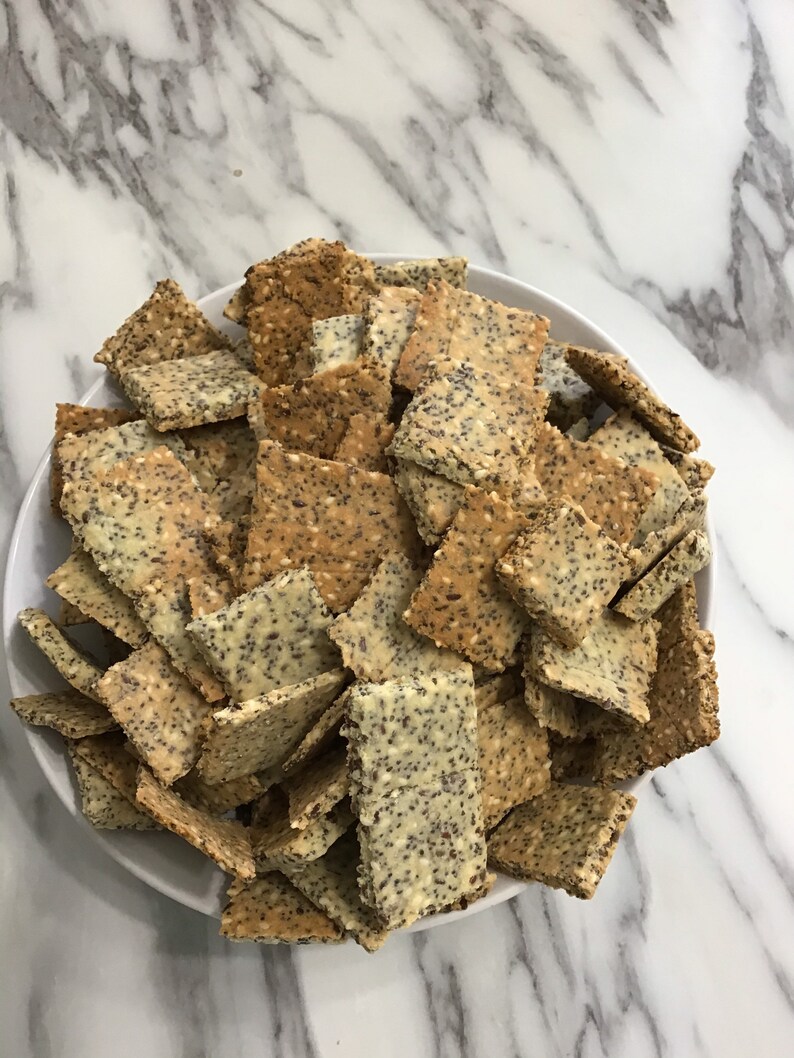May include: A white bowl filled with a variety of crackers. The crackers are rectangular and have a light brown color with a sprinkle of poppy seeds on top.