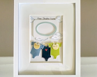 May include: A framed artwork featuring three baby onesies in blue, navy, and green, each with a tag. The artwork includes a space for a photo and the name "Adam Johnathon Endrie". The onesies have a heart and the dates "April 3, 2019" and "Nov. 11th".