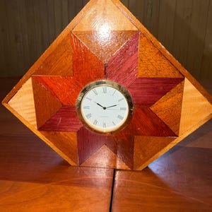 May include: A diamond-shaped wooden clock with a starburst design. The clock face is round with Roman numerals and gold trim. The wood is a mix of light and dark brown, red, and orange tones.