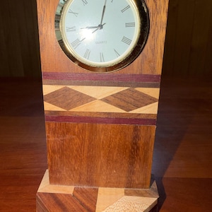 May include: A wooden quartz clock with a gold-colored bezel and Roman numeral hour markers. The clock face is white. The wooden clock has a geometric design with diamond and rectangular patterns in shades of brown and red.