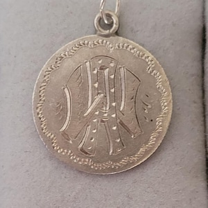 May include: Silver pendant with a detailed engraved design of a person wearing a coat. The pendant has a rope-like border around the edge.