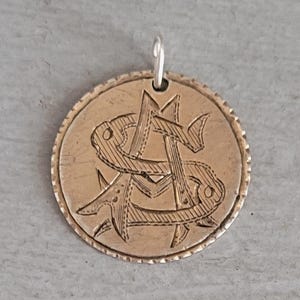 May include: A circular gold-toned metal pendant featuring an engraved monogram of the letters A and S intertwined. The pendant has a textured, scalloped edge and includes a small silver-toned metal jump ring at the top for attachment to a chain.