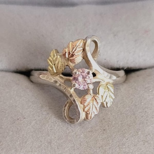 May include: A silver ring with a pink gemstone center, surrounded by gold and rose gold leaf accents. The ring has a delicate, nature-inspired design, perfect for everyday wear. The ring is a size 7.