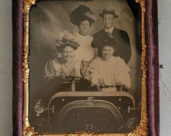 Può includere: Una fotografia vintage in bianco e nero di quattro persone in un'auto. Le persone sono vestite in abiti formali e sorridono. L'auto è un modello vintage con una grande griglia del radiatore. La foto è incorniciata in una cornice dorata con un passepartout in velluto viola.