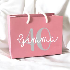 May include: A pink gift bag with white rope handles. The bag features the number "40" in gray and the name "Gemma" in a white script font. The bag is sitting on a white surface.