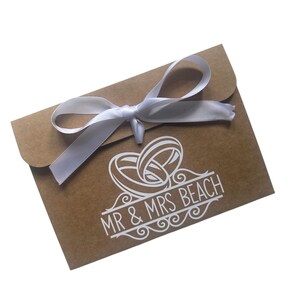 May include: A rectangular brown kraft paper envelope featuring a white satin ribbon tied in a bow at the top flap. The front of the envelope displays a white graphic of two interlocking wedding rings above the text MR & MRS BEACH.