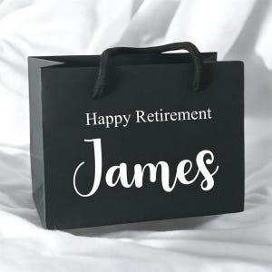 May include: A black gift bag with the words "Happy Retirement James" in white lettering. The bag has black rope handles and is on a white surface. The bag is square and has a glossy finish.
