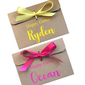 K&ouml;nnte beinhalten: Zwei Geschenk-Umschl&auml;ge aus Kraftpapier mit gelben und rosa B&auml;ndern. Der gelbe Umschlag tr&auml;gt den Text "Happy Easter Ryden". Der rosa Umschlag tr&auml;gt den Text "Happy Easter Ocean".