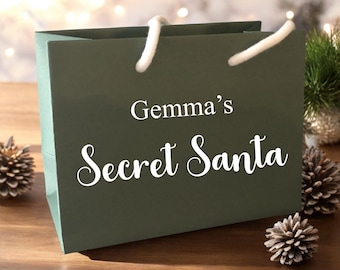 May include: A dark green gift bag with white rope handles. The text "Gemma's Secret Santa" is printed in white script on the front of the bag. Pine cones and greenery are in the background, suggesting a holiday theme.