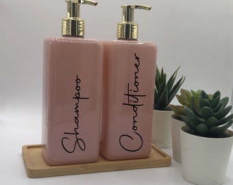 Peut inclure: Deux distributeurs de savon rectangulaires rose clair avec des pompes dorées. Les flacons portent l'inscription Shampoo et Conditioner en écriture cursive noire. Ils sont posés sur un plateau en bois rectangulaire près de deux plantes succulentes.