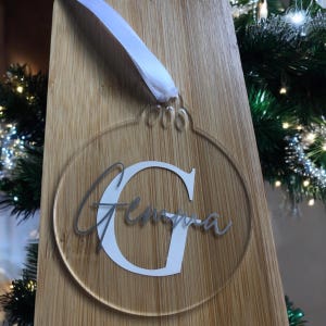 Puede incluir: Un adorno navideño de acrílico transparente con una letra G blanca y el nombre "Gemma" escrito en purpurina plateada. El adorno está colgado en una superficie de madera.