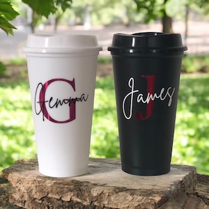 Può includere: Due tazze da viaggio personalizzate, una bianca con il nome "Gemma" in lettere rosa e una nera con il nome "James" in lettere bianche.