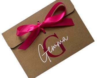 May include: A brown paper envelope tied with a bright pink satin ribbon. The envelope has the name "Gemma" printed on it in a cursive font, with a large letter "G" in a matching pink color.