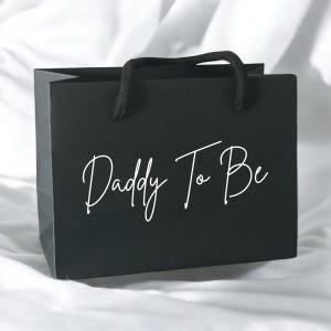 May include: A matte black gift bag featuring the white cursive text Daddy To Be printed on the front. The bag includes two black rope handles attached to the top edge, providing a simple and functional design for carrying small items or gifts.