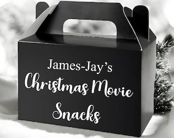 May include: A black gift box with a handle, featuring the text "James-Jay's Christmas Movie Snacks" in white script. The box has a rectangular shape and a glossy finish. A small evergreen branch is visible on the right.