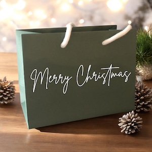 May include: A dark green gift bag with white rope handles and the words "Merry Christmas" in elegant script. The bag is on a wooden surface, with pine cones and greenery in the background, suggesting a festive holiday theme.